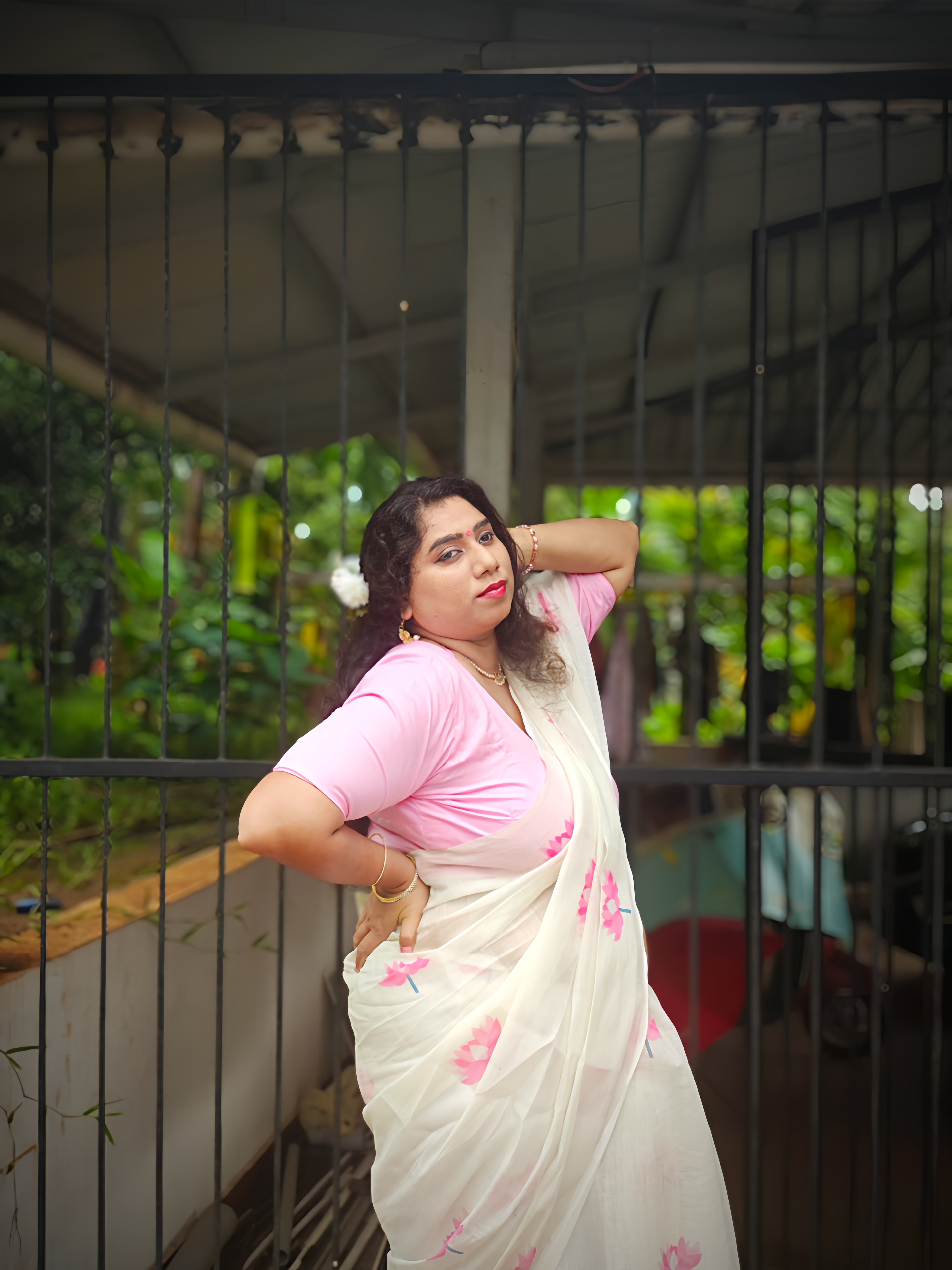 Woman wearing a soft cotton linen white saree with pink lotus floral print and matching pink blouse, posing near a black gate. Lightweight cotton linen saree for festive, office and casual wear by Vyra Fashion.