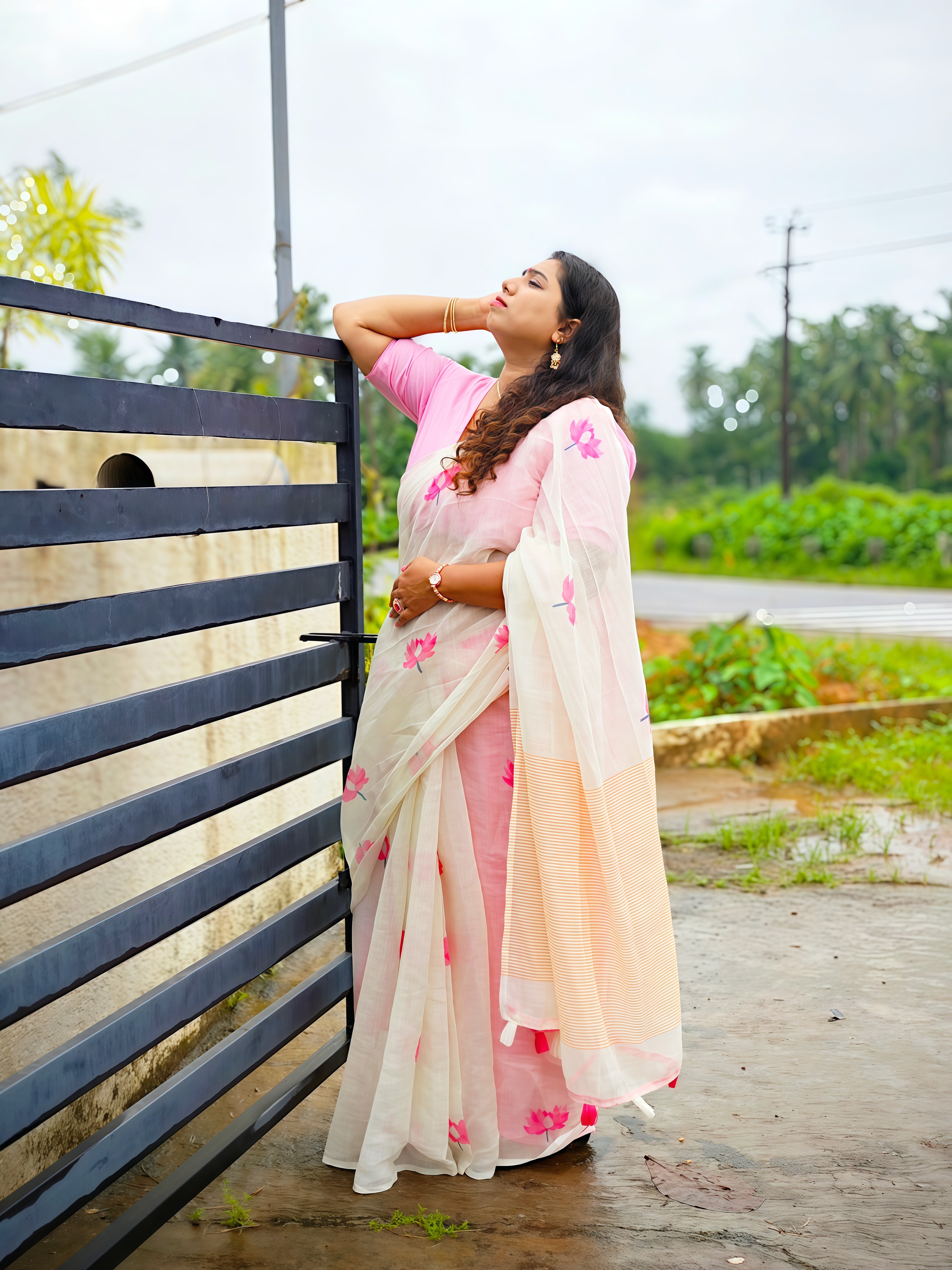 Woman wearing a soft cotton linen white saree with pink lotus floral print and matching pink blouse, posing outdoors. Lightweight festive, office and casual wear by Vyra Fashion.